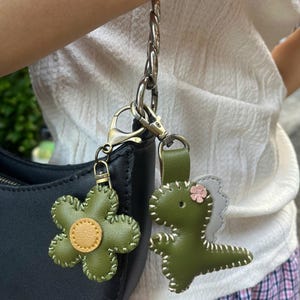 May include: Two green leather keyrings: a flower with a yellow centre and a dinosaur with a pink flower accent. Both keyrings are attached to a silver chain and a black handbag.