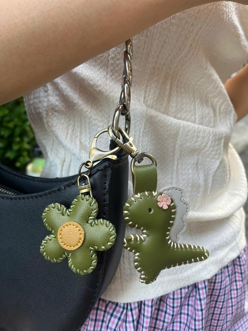 May include: Two green leather keyrings: a flower with a yellow centre and a dinosaur with a pink flower accent. Both keyrings are attached to a silver chain and a black handbag.