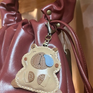 May include: A beige leather-like capybara keychain featuring a brown snout, a blue teardrop accent, and visible cream-colored stitching along the edges. The charm is attached to a bronze-toned metal lobster clasp and hangs from a dark red handbag.