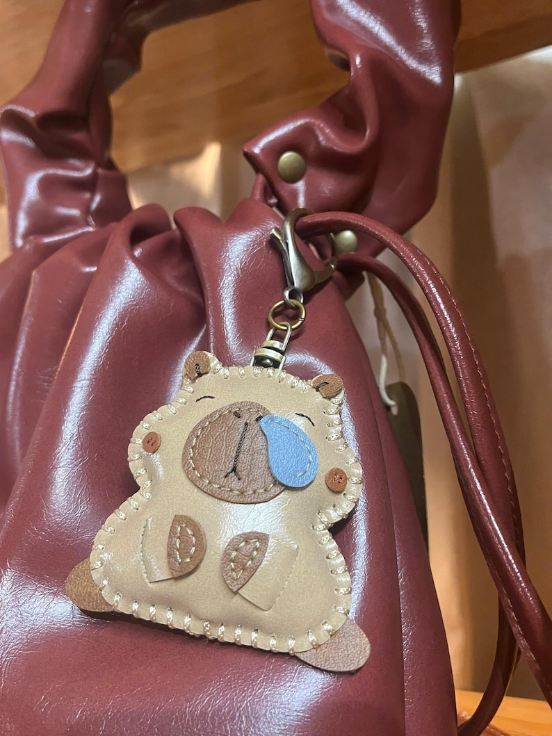 May include: A beige leather-like capybara keychain featuring a brown snout, a blue teardrop accent, and visible cream-colored stitching along the edges. The charm is attached to a bronze-toned metal lobster clasp and hangs from a dark red handbag.