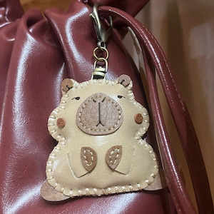 May include: A beige leather-like keychain shaped like a capybara with dark brown stitched details, a rounded snout, and small ears. The charm is attached to a dark metal lobster clasp and hangs against a dark maroon, smooth, drawstring-style bag.