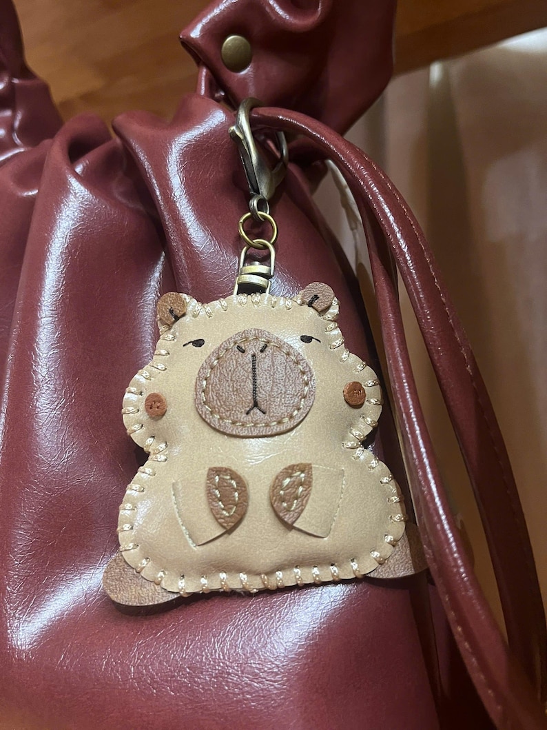 May include: A beige leather-like keychain shaped like a capybara with dark brown stitched details, a rounded snout, and small ears. The charm is attached to a dark metal lobster clasp and hangs against a dark maroon, smooth, drawstring-style bag.