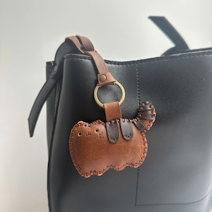 May include: A brown leather cat-shaped keyring with stitched detailing and a small tail, attached to a black handbag with a brown leather strap and a gold ring.