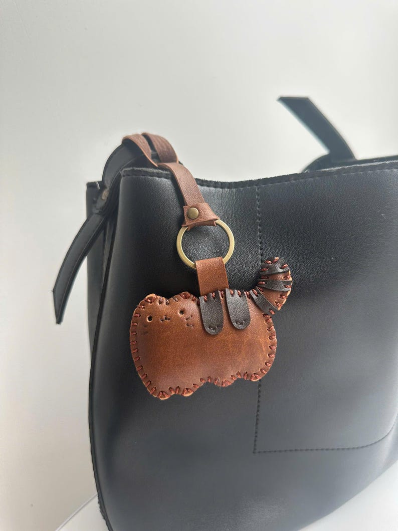 May include: A brown leather cat-shaped keyring with stitched detailing and a small tail, attached to a black handbag with a brown leather strap and a gold ring.