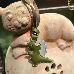 May include: A green leather dinosaur keyring with a pink flower and grey wings. The keyring has a green strap and a silver clasp. The dinosaur is stitched with gold thread. The keyring is hanging on a terracotta cat statue.