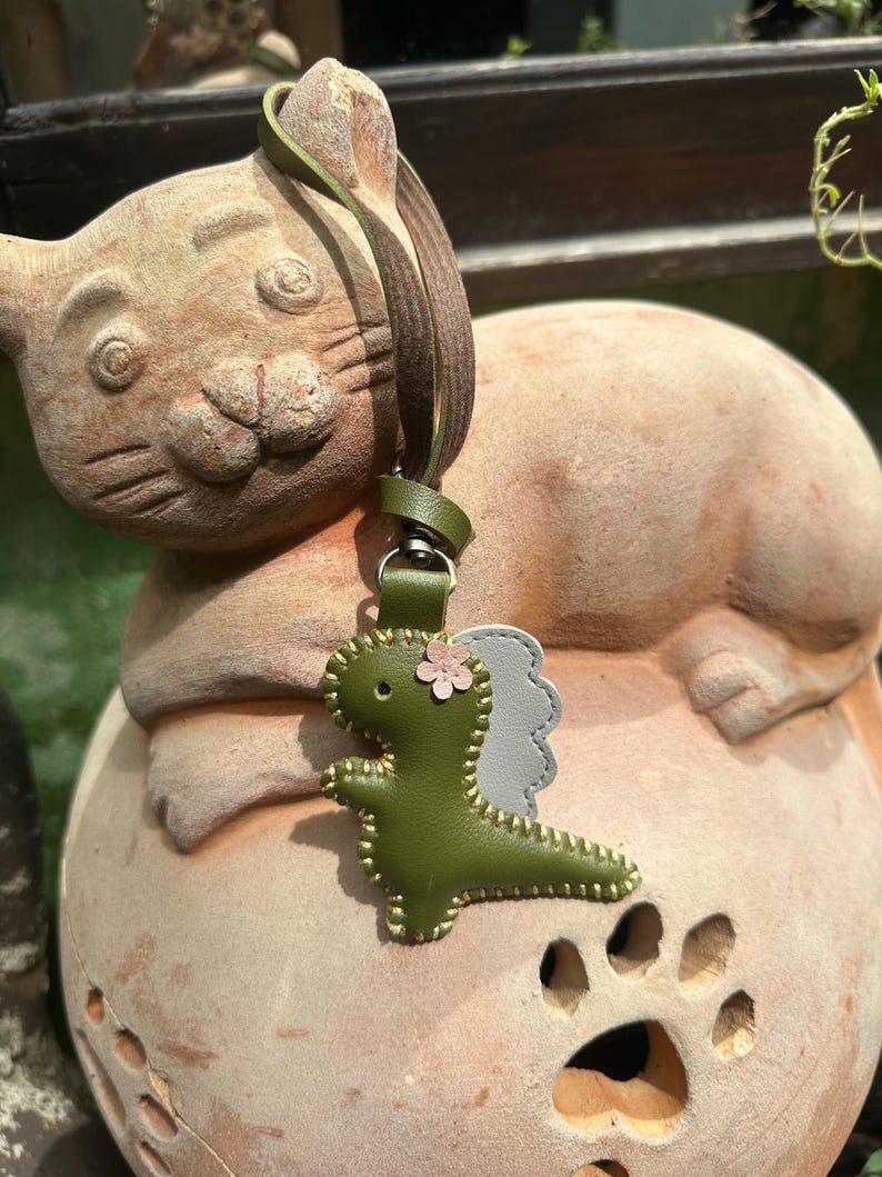 May include: A green leather dinosaur keyring with a pink flower and grey wings. The keyring has a green strap and a silver clasp. The dinosaur is stitched with gold thread. The keyring is hanging on a terracotta cat statue.