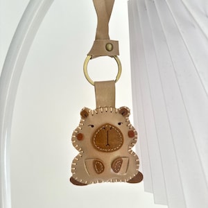 May include: A beige leather keychain shaped like a capybara, featuring a dark brown snout, stitched edges, and a brass-colored metal ring attached to a leather strap. The design includes small brown circular accents on the cheeks and paws.