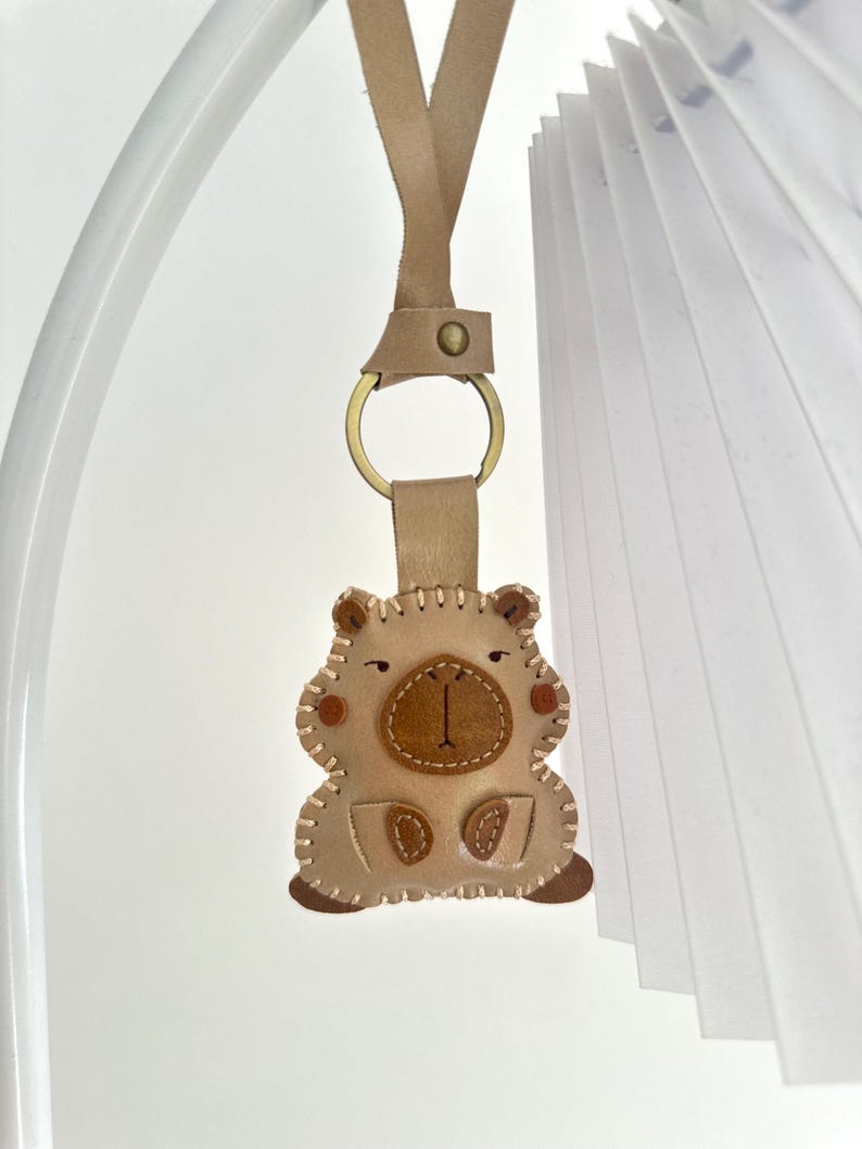 May include: A beige leather keychain shaped like a capybara, featuring a dark brown snout, stitched edges, and a brass-colored metal ring attached to a leather strap. The design includes small brown circular accents on the cheeks and paws.