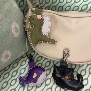 May include: Three leather keyrings: a green dinosaur with a pink flower, a purple whale with a pink heart, and a black cat with yellow eyes. Each keyring has a metal clasp for easy attachment. The keyrings are on a green and white patterned background.