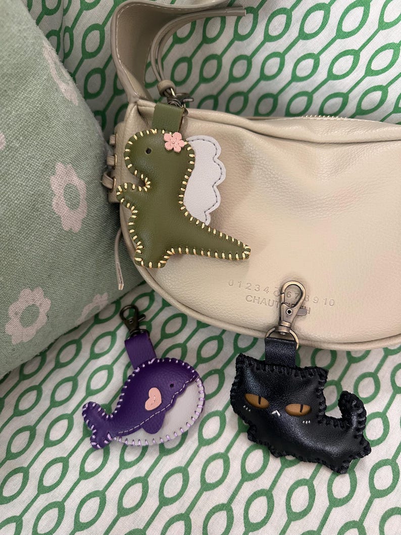 May include: Three leather keyrings: a green dinosaur with a pink flower, a purple whale with a pink heart, and a black cat with yellow eyes. Each keyring has a metal clasp for easy attachment. The keyrings are on a green and white patterned background.