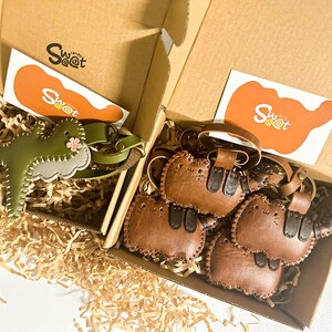 May include: Two open cardboard boxes filled with decorative paper and leather keyrings. One box contains a green dinosaur keyring with a pink flower, and the other has several brown cat-shaped keyrings. Each box has a card with the text "Sweet".