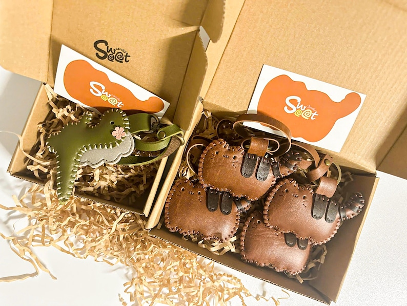 May include: Two open cardboard boxes filled with decorative paper and leather keyrings. One box contains a green dinosaur keyring with a pink flower, and the other has several brown cat-shaped keyrings. Each box has a card with the text "Sweet".