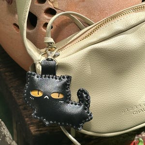 May include: A black leather cat-shaped keyring with yellow eyes and white details. The keyring is attached to a pale green leather bag with gold-coloured hardware. The cat charm is stitched with a contrasting thread.
