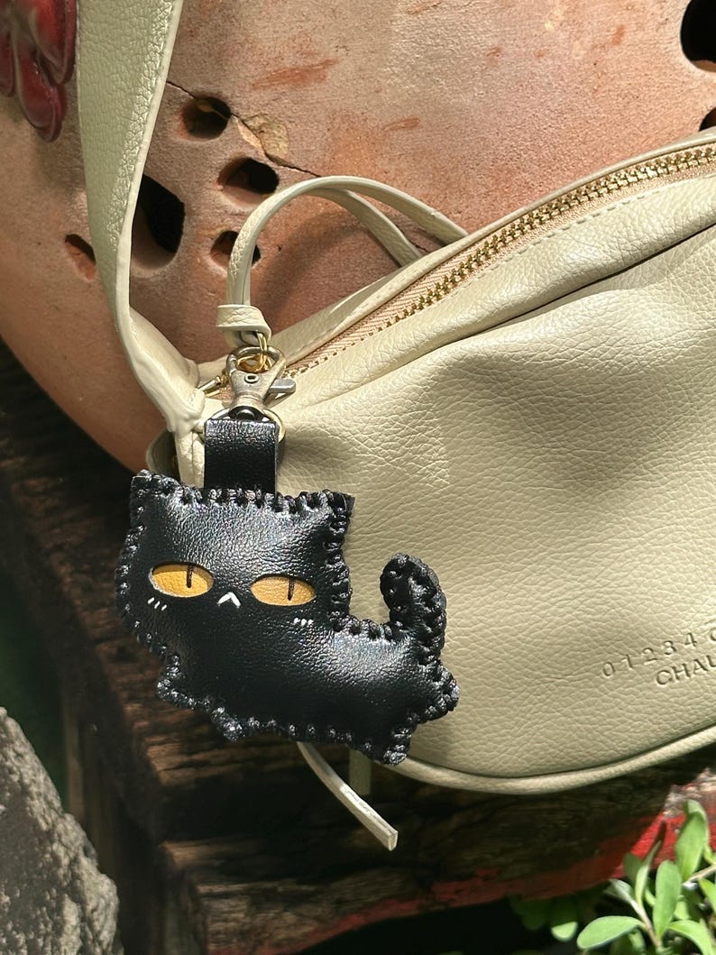 May include: A black leather cat-shaped keyring with yellow eyes and white details. The keyring is attached to a pale green leather bag with gold-coloured hardware. The cat charm is stitched with a contrasting thread.