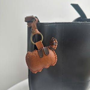 May include: A brown leather cat-shaped keyring with a stitched border and a tail, attached to a black tote bag. The keyring has a gold-coloured ring and a brown leather strap.