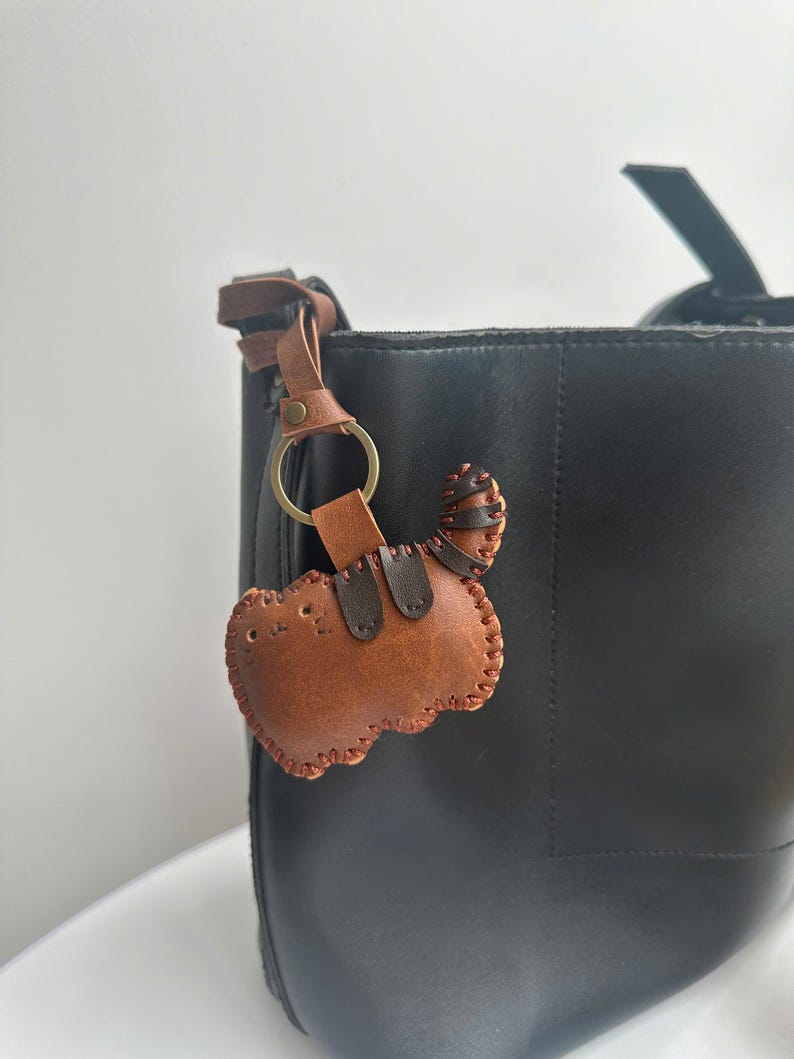 May include: A brown leather cat-shaped keyring with a stitched border and a tail, attached to a black tote bag. The keyring has a gold-coloured ring and a brown leather strap.