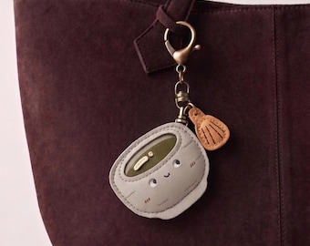 Könnte beinhalten: Ein hellgrauer Schlüsselanhänger aus Leder in Form einer Schale mit lächelndem Gesicht, mit einem dunkelgrünen Innendetail und einem kleinen beigen muschelförmigen Anhänger an einem bronzefarbenen Karabinerhaken und Ring.