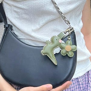 May include: Black crescent-shaped handbag with a silver chain strap. The bag features two leather charms: a green dinosaur and a green flower. The dinosaur charm has white stitching and a small pink flower.