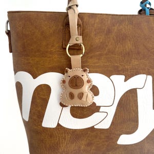 May include: A brown faux leather tote bag with a large white lettered design and a beige leather keychain shaped like a small animal with stitched details, attached to the handle by a gold metal ring and a light tan leather strap.