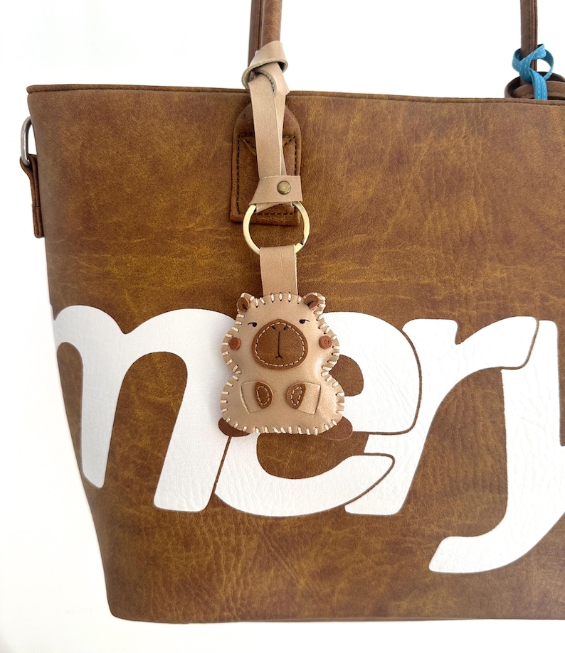 May include: A brown faux leather tote bag with a large white lettered design and a beige leather keychain shaped like a small animal with stitched details, attached to the handle by a gold metal ring and a light tan leather strap.