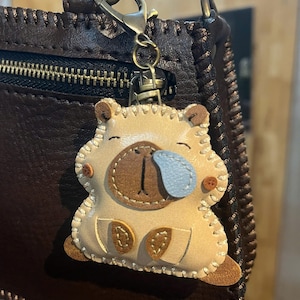 May include: A beige leather capybara keychain featuring a light blue teardrop detail, dark brown stitched accents, and a bronze-toned metal lobster clasp. The charm is attached to a dark brown textured leather bag with visible stitching along the edges.