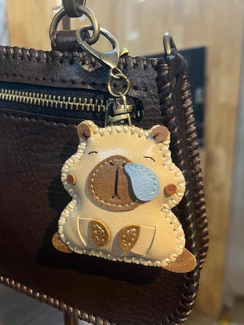 May include: A beige leather capybara keychain featuring a light blue teardrop detail, dark brown stitched accents, and a bronze-toned metal lobster clasp. The charm is attached to a dark brown textured leather bag with visible stitching along the edges.