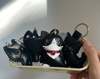 May include: A collection of black cat-shaped keychains made of stitched leather. The central cat features a white face mask and a red collar with a small circular charm. Each keychain includes a metal ring attachment for keys or bags.