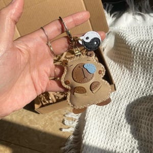May include: A tan leather-like keychain shaped like a stylized bear with a dark brown snout, a light blue nose, and small brown paws. The keychain features a metal ring attached to a small, circular white and black yin-yang charm.