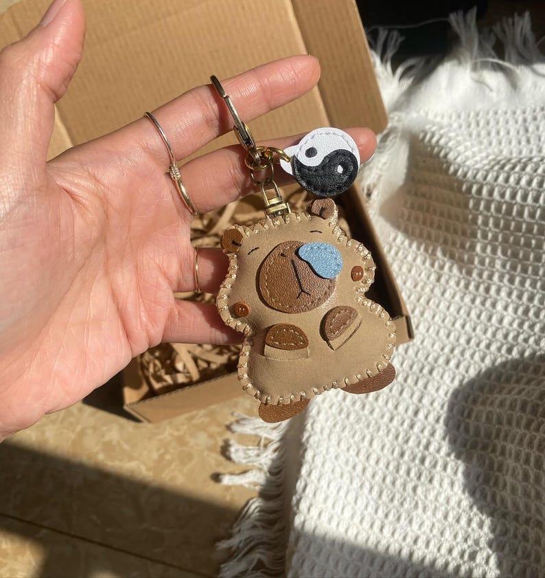 May include: A tan leather-like keychain shaped like a stylized bear with a dark brown snout, a light blue nose, and small brown paws. The keychain features a metal ring attached to a small, circular white and black yin-yang charm.