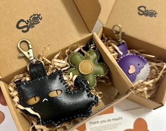May include: Three leather keychains in cardboard boxes. One black cat keychain with gold eyes, one green flower keychain, and one purple whale keychain. The boxes have the word "Sweet" printed on them.