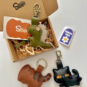 May include: A gift box containing three leather keyrings: a green dinosaur, a brown dog, and a black cat. The dinosaur keyring has a pink flower. The box includes a sticker that says "Thank You" and a Sweet candy logo.