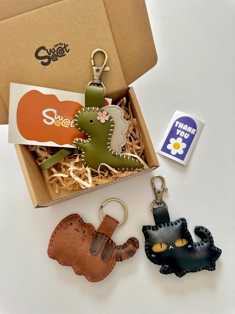 May include: A gift box containing three leather keyrings: a green dinosaur, a brown dog, and a black cat. The dinosaur keyring has a pink flower. The box includes a sticker that says "Thank You" and a Sweet candy logo.