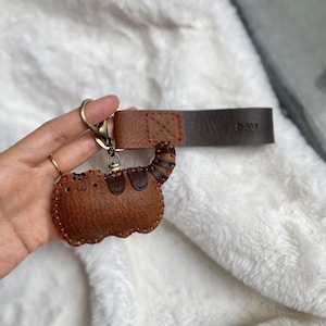 May include: A brown leather cat-shaped keyring with stitched detailing. It has a metal clasp and a leather strap with the text "DAU". The cat's tail features a striped pattern.