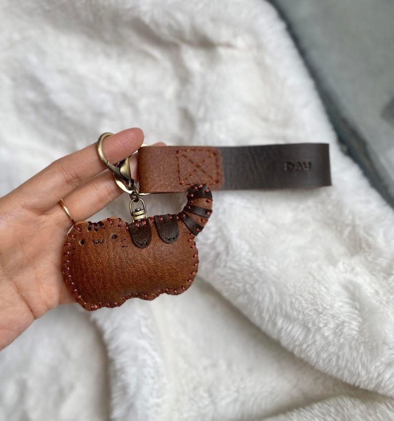 May include: A brown leather cat-shaped keyring with stitched detailing. It has a metal clasp and a leather strap with the text "DAU". The cat's tail features a striped pattern.