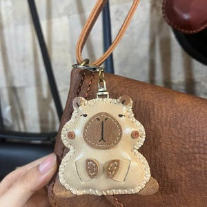 May include: A beige leather keychain shaped like a stylized capybara with a dark brown snout, small ears, and stitched edges. The charm is attached to a tan leather strap with a metal lobster clasp, hanging against a brown leather bag.