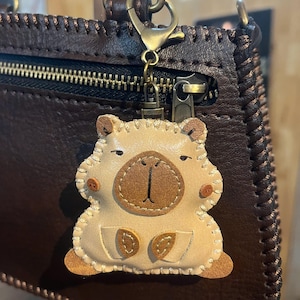 May include: A beige leather animal-shaped keychain with stitched edges, a dark brown nose patch, and small brown feet, attached to a dark brown leather bag by a bronze-colored metal lobster clasp and a short chain link connector.