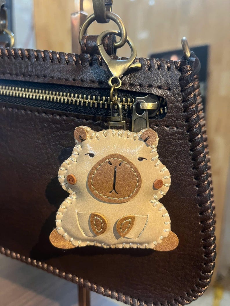 May include: A beige leather animal-shaped keychain with stitched edges, a dark brown nose patch, and small brown feet, attached to a dark brown leather bag by a bronze-colored metal lobster clasp and a short chain link connector.