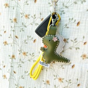 May include: A green leather dinosaur keyring with a yellow strap and a silver clasp. The dinosaur has a pink flower and white stitching. A black car key is also attached to the keyring.