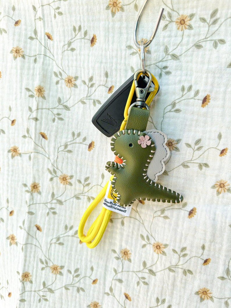 May include: A green leather dinosaur keyring with a yellow strap and a silver clasp. The dinosaur has a pink flower and white stitching. A black car key is also attached to the keyring.