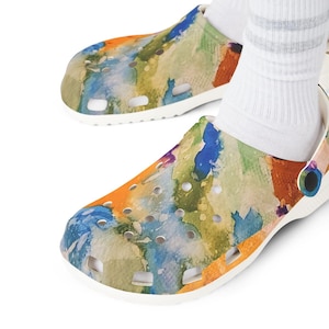 Op de afbeelding: Witte klompen met een kleurrijk abstract aquarelontwerp. De klompen hebben een mix van oranje, blauwe, groene en gele tinten. De schoenen hebben meerdere ventilatiegaten en een witte band. De klompen zijn ongeveer 25 cm lang.