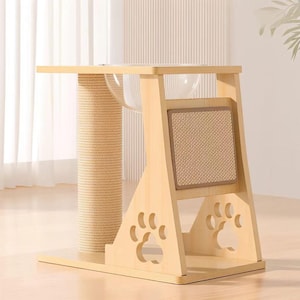 Puede incluir: Un árbol para gatos de madera clara con un cuenco transparente, un poste rascador y una almohadilla rascadora. La estructura presenta recortes en forma de huellas de pata y una plataforma. El poste rascador está envuelto en cuerda de sisal de color natural.