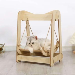 Op de afbeelding: Een lichtgekleurde houten kattenzwaai met een grijs-witte kat die op het platform van de zwaai rust. De zwaai heeft een rechthoekige basis en een gebogen bovenkant, met touwsteunen. De kat ligt en kijkt naar de camera.