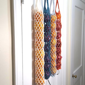May include: Five crocheted produce bags in white, teal, gray, and orange, filled with oranges and other fruits. The bags hang from a white door, showcasing a handmade, eco-friendly storage solution for fruits and vegetables.