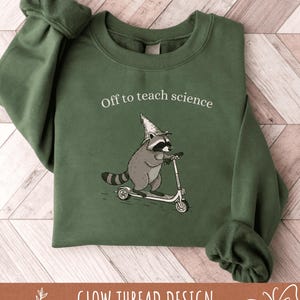 May include: Green sweatshirt with the text "Off to teach science." The graphic features a raccoon wearing a wizard hat and riding a scooter. The bottom of the sweatshirt has the text "GLOW THREAD DESIGN."