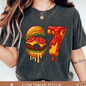 May include: Dark gray t-shirt featuring a graphic of a burger and fries forming the number 67. The burger has tomatoes, cheese, and lettuce. The fries are yellow with red ketchup dripping down. The text "GLOW THREAD DESIGN" is at the bottom.