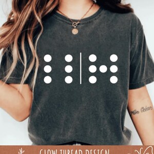 May include: Dark gray t-shirt with a white braille design. The design consists of six white dots and a vertical line. The text "GLOW THREAD DESIGN" is at the bottom of the image.