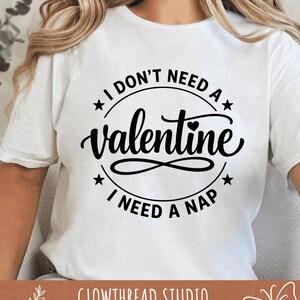 May include: White t-shirt with a black circular design that reads "I DON'T NEED A Valentine, I NEED A NAP." The word "Valentine" is in a cursive font, and the design includes stars and a heart. The brand name "GLOWTHREAD STUDIO" is at the bottom.