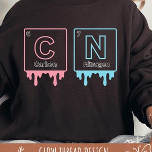 May include: A dark brown sweatshirt features a graphic design of the periodic table elements Carbon and Nitrogen. The pink and blue squares have a dripping paint effect. The text "Carbon" and "Nitrogen" are below the letters. The words "Glow Thread Design" are at the bottom.