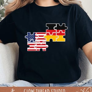 Puede incluir: Camiseta negra con un diseño de rompecabezas que representa las banderas estadounidense y alemana. La bandera estadounidense está representada en piezas de rompecabezas rojas, blancas y azules, mientras que la bandera alemana está en piezas negras, rojas y amarillas. El texto "GLOW THREAD STUDIO" está en la parte inferior.