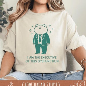 May include: Cream-coloured t-shirt with a teal cartoon frog in a suit and the text "I AM THE EXECUTIVE OF THIS DYSFUNCTION." The shirt also has the text "GLOWTHREAD STUDIO" at the bottom. The frog is surrounded by four star shapes.